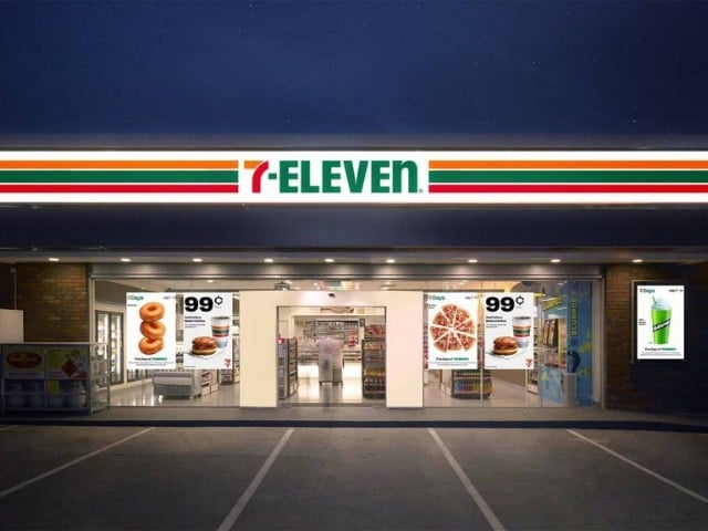 Night view of a 7-Eleven convenience store with bright signage and product posters at the entrance.