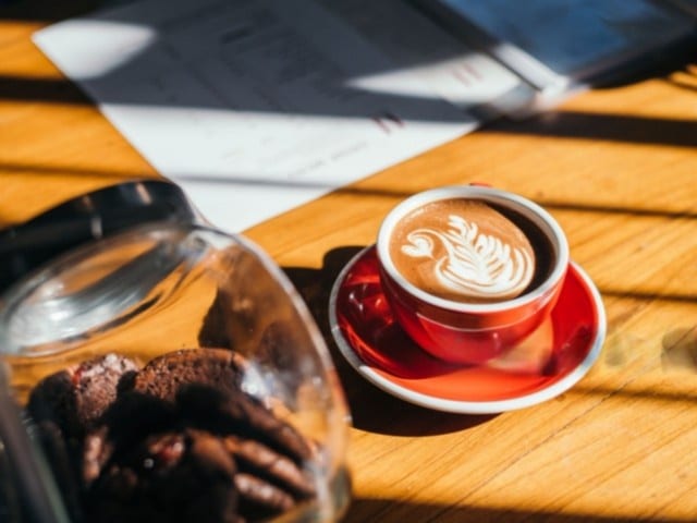 A cup of coffee with latte art on a wooden café table beside a jar of cookies.